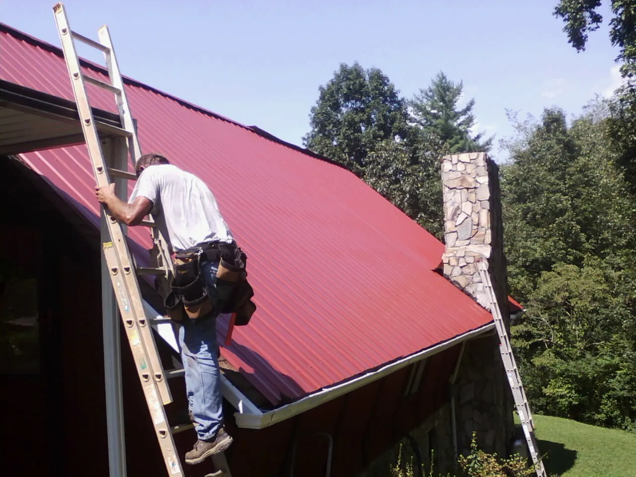 Roofer on a ladder beside a red metal roof and stone chimney — Soffit & Fascia Repair by Roofing Brothers in Davenport